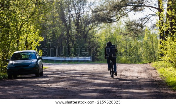 Male cyclist rides on the road in the park, forest\
on a sunny day. There is a car on the side of the road. Choice\
between bike and car.