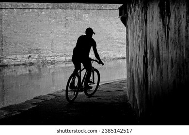 A male cyclist is silhouetted against a brick wall as he rides beneath a bridge along a narrow canal, Huddersfield, Yorkshire, England, UK.
