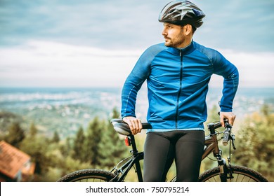 A Male Cyclist Stands On A Hill, Near The City.