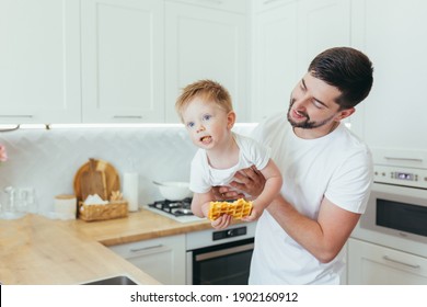 Male Dad Holding A Little Son In His Arms And Eating, Cooking At Home In The Kitchen