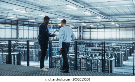 Male Data Center System Administrators and African American IT Specialists Talk, Use Laptop. Information Technology Engineers work on Cyber Security Network Protection in Cloud Computing Server Farm. - Powered by Shutterstock