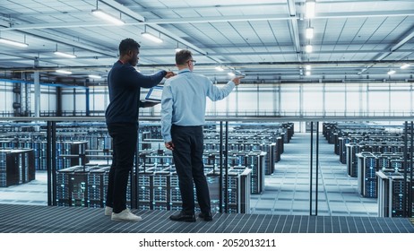 Male Data Center System Administrators And African American IT Specialists Talk, Use Laptop. Information Technology Engineers Work On Cyber Security Network Protection In Cloud Computing Server Farm.