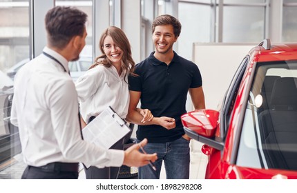 Male dealer with clipboard gesticulating and speaking with happy young man and woman near new vehicle while working in car showroom