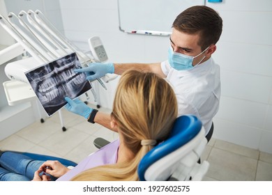 Male Dental Practitioner Wearing Hospital Mask Turning To A Lady In Medical Chair And Talking About X-ray Diagram