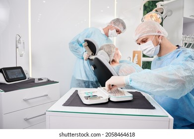 Male Dentist Choosing Dental Implants, Sitting At The Table With Dental Implant Kit While Woman Lying In Dental Chair. Concept Of Dentistry, Stomatology And Dental Care.