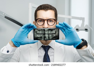 Male dentist cover his mouth with a smartphone with X-ray of his teeth on a screen of device. Modern orthodontist in dental clinic concept - Powered by Shutterstock