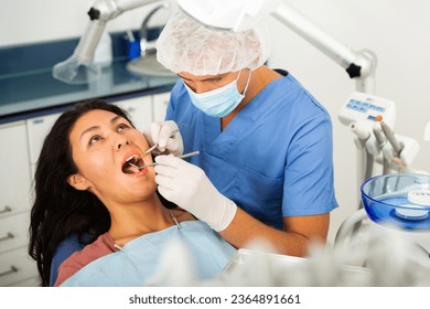 Male dentist in face mask inspects asian female patient teeth with mirror and probe - Shutterstock ID 2364891661