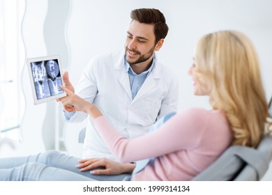 Male dentist showing female patient jaw x-ray on digital tablet, dental clinic interior. Bearded man stomatologist having conversation with lady in dental chair, pointing at gadget, side view