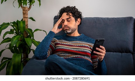 Male Depression Holding His Forehead While Having Headache On Couch Home. Adult Man Using Smart Phone And Thinking, Having Heavy Thoughts, Breake Up With Lover Or Financial Difficulties Concept