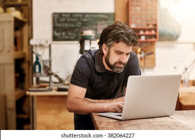 Male designer and craftsman with a rugged beard, working on his laptop at his workbench, in his studio workshop - Powered by Shutterstock