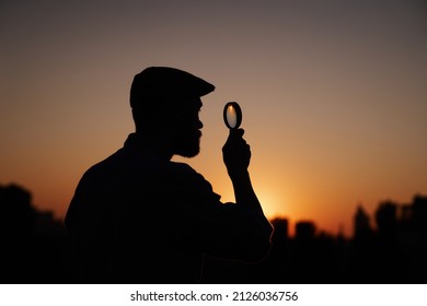 Male Detective Or Investigator Silhouette Portrait In Peaked Hat Looking With Magnifying At Side. Man Searching Using Loupe With Sun On Background And Urban View. Investigation Or Inspection Concept