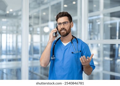 Male doctor in blue scrubs with stethoscope talking on phone inside bright, modern medical facility. Confident medical professional illustrating healthcare, communication, and technology concepts. - Powered by Shutterstock