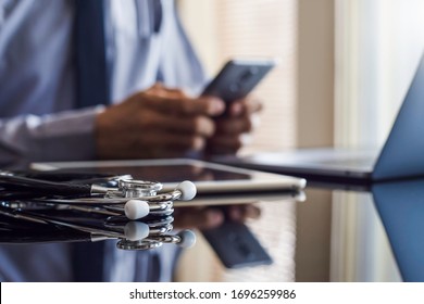 Male doctor in blue shirt with tie, hand holding and using mobile smart phone with laptop compute, medical stethoscope and digital tablet on the desk. Medical online networking, telehealth concept.