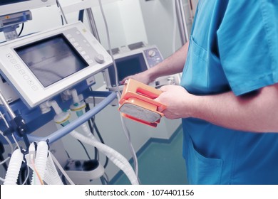 Male Doctor With Defibrillator Electrodes In His Hand Sets The Electric Discharge Parameters In Front Of The Life Support Monitor.