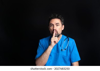 A Male Doctor Dressed In A Blue Hospital Uniform, Dark And Bearded, Wearing A Stethoscope Around His Neck, Asks For Silence With His Index Finger Over His Lips. A Black Background And Copy Space Area 