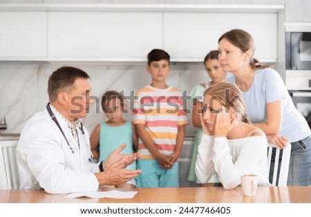 Male doctor examinating teen girl sitting at home kitchen on background of her mother and siblings