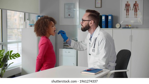 Male Doctor Giving African Little Girl Checkup In Clinic Office. Man Pediatrician Examining Child Throat With Wooden Stick In Hospital Office. Healthcare And Medicine Concept