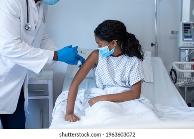 Male Doctor Giving Covid Vaccination To Sick Mixed Race Girl In Face Mask Sitting Up In Hospital Bed. Medicine, Health And Healthcare Services During Coronavirus Covid 19 Pandemic.