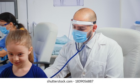 Male Doctor Giving Girl Checkup In Doctor Office Using Stethoscope During Coronavirus. Physician Specialist In Medicine Providing Health Care Services Consultation Treatment Examination In Hospital