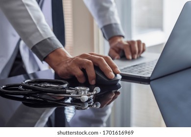 Male Doctor Hand Using Wireless Mouse, Browsing Medical Information On Laptop Computer With Stethoscope On The Desk. Medic Tech, Telehealth , Online Medical, Teleconference Or Telemedicine Concept. 