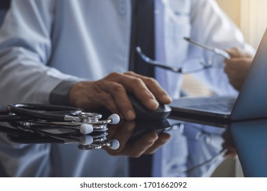 Male Doctor Hand Using Wireless Mouse, Browsing Medical Information On Laptop Computer With Stethoscope On The Desk. Medic Tech, Telehealth , Online Medical, Teleconference Or Telemedicine Concept. 