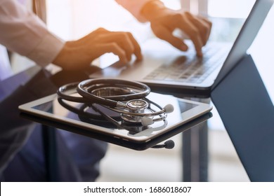 Male Doctor Hands Typing On Laptop Computer Keyboard, Search Medical Information With Digital Tablet Pc And Medical Stethoscope On The Desk At Office. Online Medical,medic Tech, Emr, Ehr Concept.  