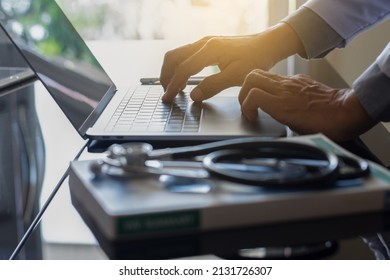 Male Doctor Hands Typing On Laptop Computer Keyboard With Textbook And Medical Stethoscope On The Desk At Office. Online Medical,medic Tech, Emr, Ehr Concept.  