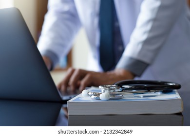 Male doctor hands typing on laptop computer keyboard with textbook and medical stethoscope on the desk at office. Online medical,medic tech, emr, ehr concept.  