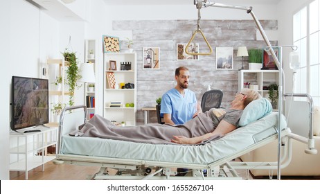 Male Doctor Having A Conversation With Ill Old Lady Lying In Hospital Bed In A Nursing Home, Medium Shot.