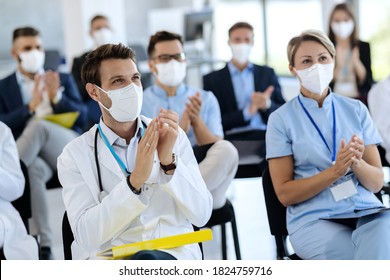 Male doctor and his colleagues wearing protective face masks and clapping their hands after successful education event in convention center. 