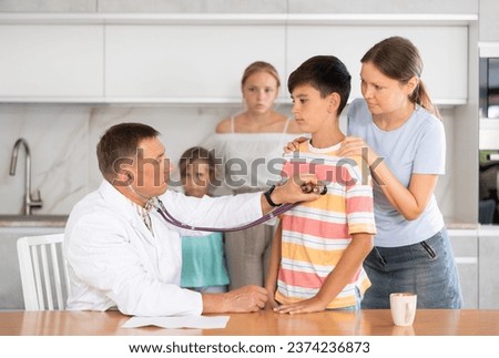 .Male doctor home visiting pre-teen boy sitting at home kitchen on background of his mother and siblings