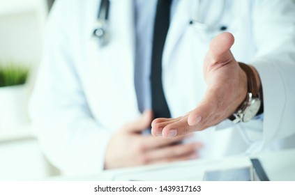 Male Doctor Making Welcome Gesture, Politely Inviting Patient To Sit Down In Medical Office. Photo With Depth Of Field. Just Hands Over The Table.