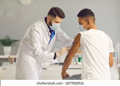 Male doctor in medical face mask giving antivirus shot to patient during vaccination campaign at the clinic. Young African-American man getting Covid-19, AIDS or flu vaccine injection at the hospital - Powered by Shutterstock