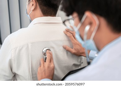 Male Doctor In Medical Face Mask Using Stethoscope To Listening Heart And Lungs From Male Patient's Back During Explaining Symptom And Giving Advice To Patient In Clinic