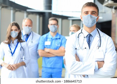 Male Doctor In Medical Mask With Group Of Happy Successful Colleagues Also In Preventive Masks.