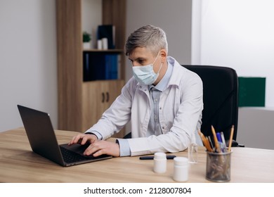 Male Doctor In Medical Mask And Uniform Working Typing On Computer. Family Therapist Consulting Patients Online Through Digital Health Platform.