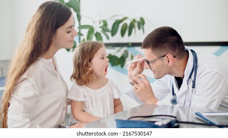 Male Doctor Pediatrician Examining An Ill Sad Kid Girl At Medical Visit With Mother In The Hospital. Male Family Doctor Examining And Consulting To Mother And Her Ill Child.