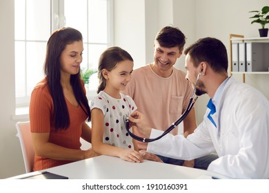 Male doctor puts a stethoscope to the little girl's chest and listens to the heartbeat and lungs. Child with mom and dad at a pediatrician. Concept of health care and pediatric medical examination.