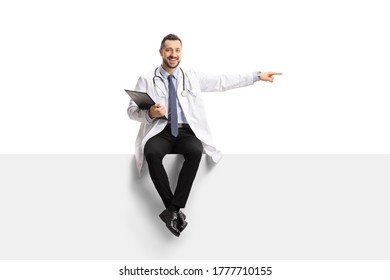 Male Doctor Sitting On A Blank Panel And Pointing To The Side Isolated On White Background