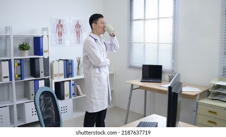 Male Doctor Standing In His Bright Office Is Looking Afar And Putting Hand Into His Pocket While Sipping Morning Coffee In Contemplation.