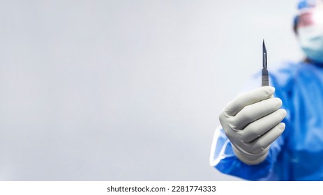 Male doctor or surgeon in blue gown holding surgical scalpel or medical knife with blur white wall background.Surgical blade for cosmetic or orthopedic surgery with space.Man wearing loupe with light. - Powered by Shutterstock