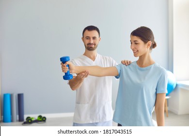 Male Doctor Teaching Happy Young Woman To Do Osteoporosis Treatment Exercise With Dumbbells In Modern Clinic. Physiotherapist Helping Female Patient During Muscle Rehabilitation Physiotherapy