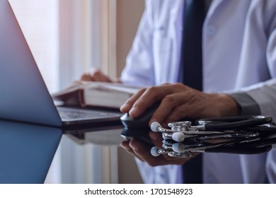 Male doctor in uniform reading book and work on laptop computer by wireless mouse with medicals stethoscope on the desk at office. Online medical technology concept.