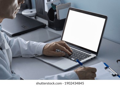 Male doctor using laptop computer with white mockup screen technology tele medicine healthcare e website ad, watching e learning medical online telehealth webinar, writing report. Over shoulder view