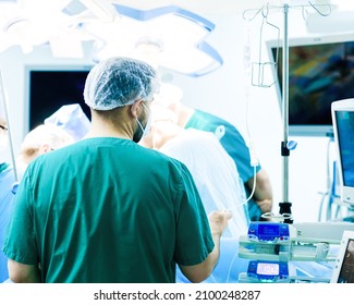 A Male Doctor Wearing A Medical Mask And Uniform Checks A Saline Drip. Back View. Background Of The Surgical Operational. 