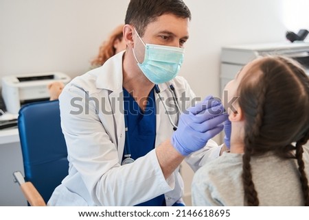 Male doctor wearing protective mask examining child girl with stetoscope at medical office