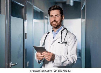 Male doctor wearing white coat stethoscope around neck, holding tablet. Confident professional standing in modern hospital hallway, smiling. Healthcare, medical professional, technology integration. - Powered by Shutterstock