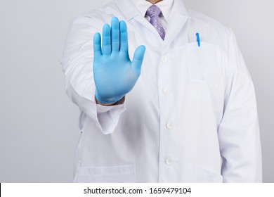 Male Doctor In A White Coat And Blue Sterile Gloves Shows A Stop Gesture With His Right Hand, White Background