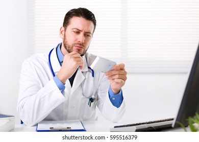 Male Doctor In White Coat With Stethoscope Over His Neck Sitting At Table Thinking On Prescription And Reading Medicine Label Of Box In His Hand