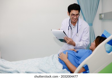 Male Doctor In White Gown Uniform Holding Chart And Talking To A Pregnant Woman Laying On Bed In Hospital Room With Care And Happy.